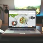 MacBook Pro displaying food blog website with salad photography, alongside cactus plant and water bottle on wooden desk