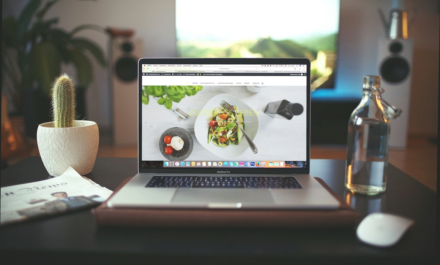 MacBook Pro displaying food blog website with salad photography, alongside cactus plant and water bottle on wooden desk